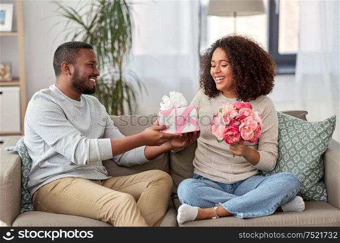 holiday, greeting and people concept - happy african american couple with bunch of flowers and gift at home. happy couple with flowers and gift at home