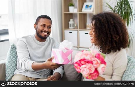 holiday, greeting and people concept - happy african american couple with bunch of flowers and gift at home. happy couple with flowers and gift at home