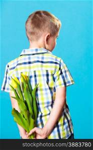 Holiday mother&#39;s day concept. Little boy has prepared surprise present for mum, flowers yellow tulips, holds it behind the back on blue