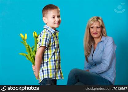 Holiday mother&#39;s day concept. Rear view little boy with bunch of yellow tulips behind back preparing nice surprise for his mother studio shot on blue