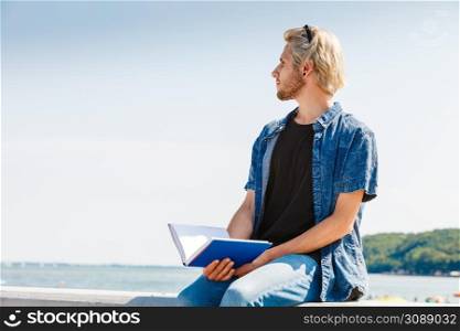 Holiday, outdoor leisure time, introvert relaxation concept. Sitting hipster man wearing jeans outfit having break from reading book outside on sunny day, sea in background. Sitting man reading book outside on sunny day
