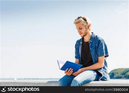 Holiday, outdoor leisure time, introvert relaxation concept. Sitting hipster man wearing jeans outfit reading book outside on sunny day, sea in background. Sitting man reading book outside on sunny day