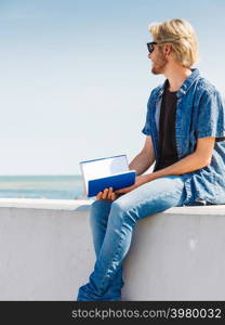 Holiday, outdoor leisure time, introvert relaxation concept. Sitting hipster man wearing jeans outfit having break from reading book outside on sunny day, sea in background. Sitting man reading book outside on sunny day