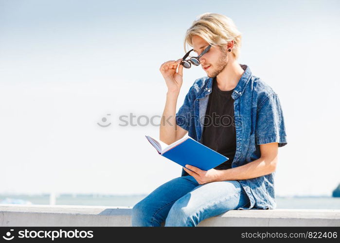 Holiday, outdoor leisure time, introvert relaxation concept. Sitting hipster man wearing jeans outfit reading book outside on sunny day, sea in background. Sitting man reading book outside on sunny day