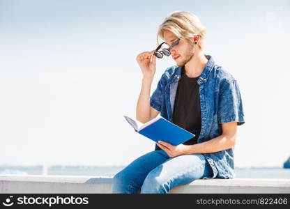 Holiday, outdoor leisure time, introvert relaxation concept. Sitting hipster man wearing jeans outfit reading book outside on sunny day, sea in background. Sitting man reading book outside on sunny day