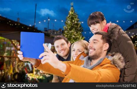 holiday, people and leisure concept - happy friends with tablet pc computer taking selfie over outdoor christmas market lights background. friends with tablet pc taking christmas selfie