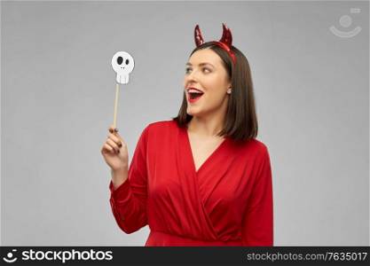 holiday, photo booth and people concept - happy smiling woman in red halloween costume of devil with horns and scull party accessory over grey background. happy woman in red halloween costume of devil