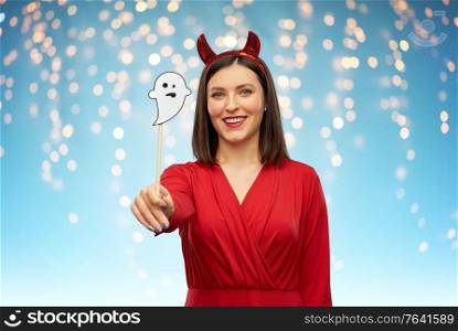 holiday, photo booth and people concept - happy smiling woman in red halloween costume of devil with horns and ghost party accessory over festive lights on blue background. happy woman in red halloween costume of devil