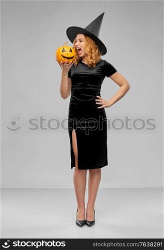holiday, theme party and people concept - angry woman in black halloween costume of witch biting jack-o-lantern pumpkin over grey background. woman in halloween costume of witch biting pumpkin