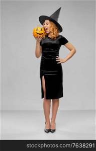 holiday, theme party and people concept - angry woman in black halloween costume of witch biting jack-o-lantern pumpkin over grey background. woman in halloween costume of witch biting pumpkin