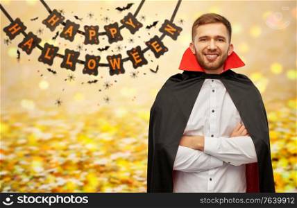 holiday, theme party and people concept - happy smiling man in halloween costume of vampire and dracula cape with arms crossed over garland string decoration and lights on background. happy man in halloween costume of vampire