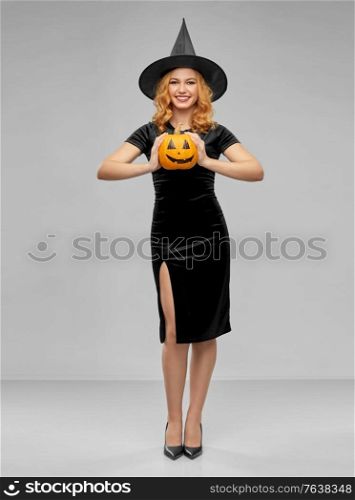 holiday, theme party and people concept - happy smiling woman in black halloween costume of witch with jack-o-lantern pumpkin over grey background. woman in halloween costume of witch with pumpkin