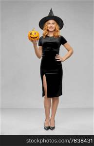 holiday, theme party and people concept - happy smiling woman in black halloween costume of witch with jack-o-lantern pumpkin over grey background. woman in halloween costume of witch with pumpkin