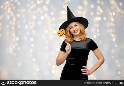 holiday, theme party and photo booth concept - happy smiling woman in black halloween costume of witch with jack-o-lantern pumpkin accessory over festive lights background. woman in halloween costume of witch with pumpkin
