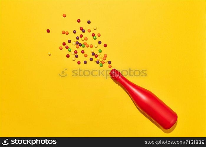 Holiday wine bottle painted red with colorful candies as a foam bubbles on an yellow background, copy space. Top view. Holiday greeting card.. Red painted champagne bottle with colorful candies.