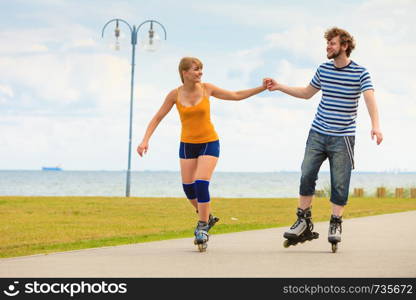 Holidays, active people and friendship concept. Young fit couple on roller skates riding outdoors on sea shore, woman and man rollerblading together on the promenade