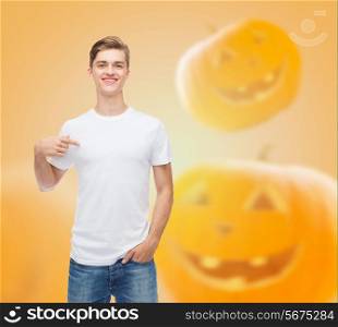 holidays, advertisement and people concept - smiling young man in blank white t-shirt pointing finger at himself over halloween pumpkins background