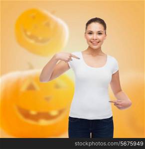 holidays, advertisement and people concept - smiling young woman in blank white t-shirt pointing fingers at herself over halloween pumpkins background