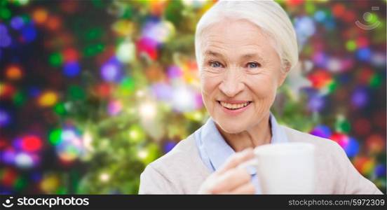 holidays, age, drink and people concept - happy smiling senior woman with cup of tea or coffee over christmas lights background