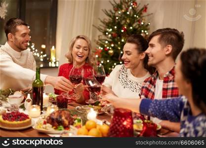 holidays and celebration concept - happy friends having christmas dinner at home, drinking red wine and clinking glasses. friends celebrating christmas and drinking wine