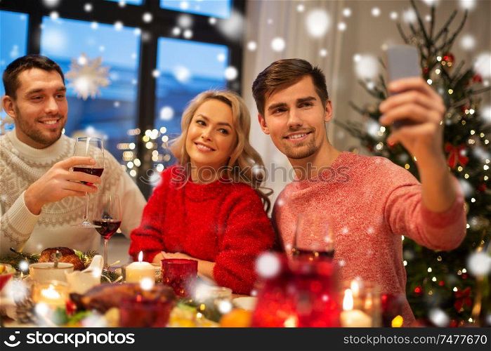 holidays and celebration concept - happy friends taking selfie by smartphone at home christmas dinner over snow. friends taking selfie at christmas dinner