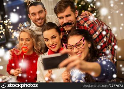 holidays and celebration concept - happy friends with party props taking selfie by smartphone at home christmas dinner over snow. friends taking selfie at christmas dinner