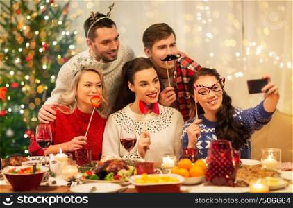 holidays and celebration concept - happy friends with party props taking selfie by smartphone at home christmas dinner. friends taking selfie at christmas dinner