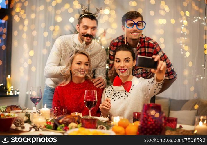 holidays and celebration concept - happy friends with party props taking selfie by smartphone at home christmas dinner. friends taking selfie at christmas dinner