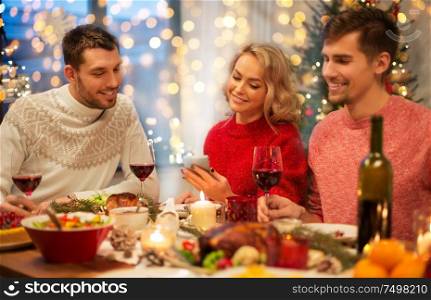 holidays and celebration concept - happy friends with smartphone having christmas dinner at home. friends with cellphone celebrate christmas at home