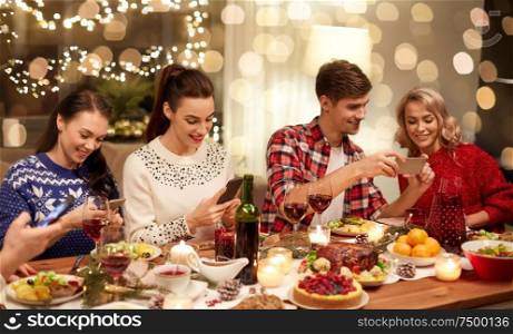 holidays and celebration concept - happy friends with smartphones having christmas dinner at home. friends with smartphones having christmas dinner