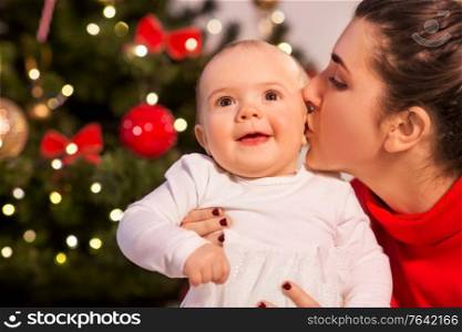 holidays and childhood concept - mother kissing happy baby daughter over christmas tree lights at home. mother kissing baby daughter over christmas tree