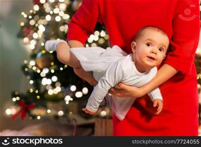 holidays and childhood concept - mother playing with baby daughter over christmas tree background at home. mother with happy baby daughter at christmas tree