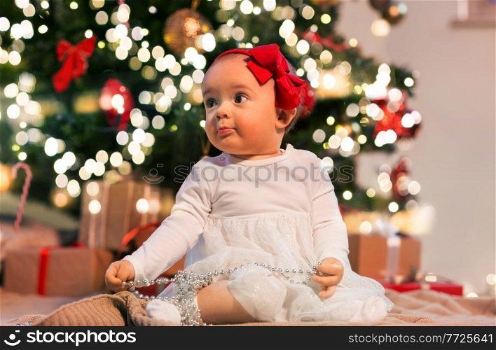 holidays and childhood concept - sweet baby girl with gifts at home over christmas tree lights. baby girl at christmas tree with gifts at home