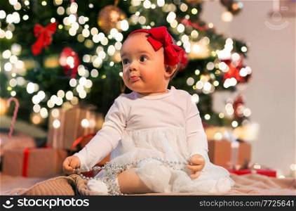 holidays and childhood concept - sweet baby girl with gifts at home over christmas tree lights. baby girl at christmas tree with gifts at home