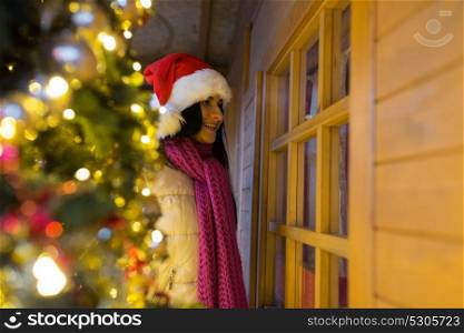 holidays and decoration concept - woman looking to santa house or shop window at christmas market. woman at santa house window at christmas market