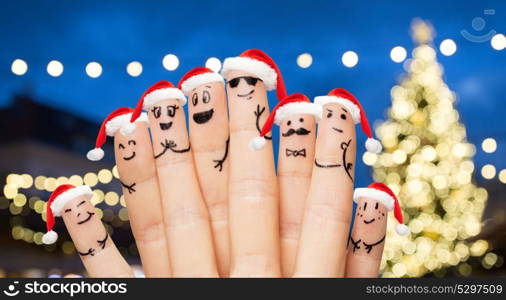 holidays and family concept - fingers with smiley faces in santa hats over christmas tree lights background. fingers in santa hats over night lights background