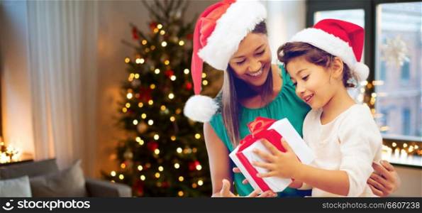 holidays and family concept - happy mother and daughter in santa hats with gift box over room with christmas tree background. happy mother and daughter with christmas gift