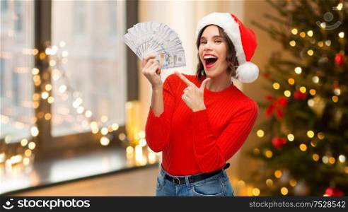 holidays and finance concept - happy smiling young woman in santa helper hat holding dollar money banknotes over christmas tree lights at home background. happy woman in santa hat with money on christmas