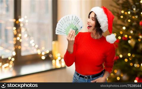 holidays and finance concept - happy smiling young woman in santa helper hat holding euro money banknotes over christmas tree lights at home background. happy woman in santa hat with money on christmas
