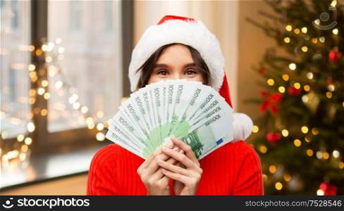 holidays and finance concept - happy young woman in santa helper hat hiding behind euro money banknotes over christmas tree lights at home background. happy woman in santa hat with money on christmas