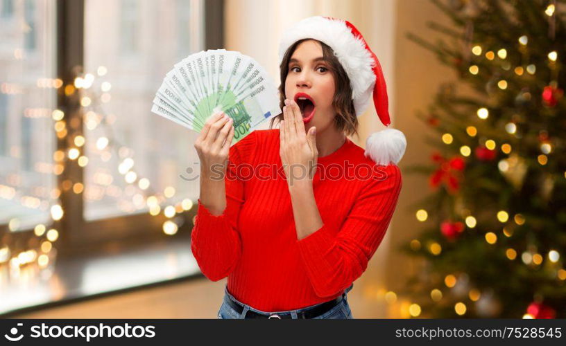 holidays and finance concept - surprised young woman in santa helper hat holding euro money banknotes over christmas tree lights at home background. surprised woman in santa hat with money