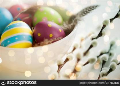 holidays and object concept - colored easter eggs with quail feathers in bowl and pussy willow branches over bokeh lighs. colored easter eggs and pussy willow branches