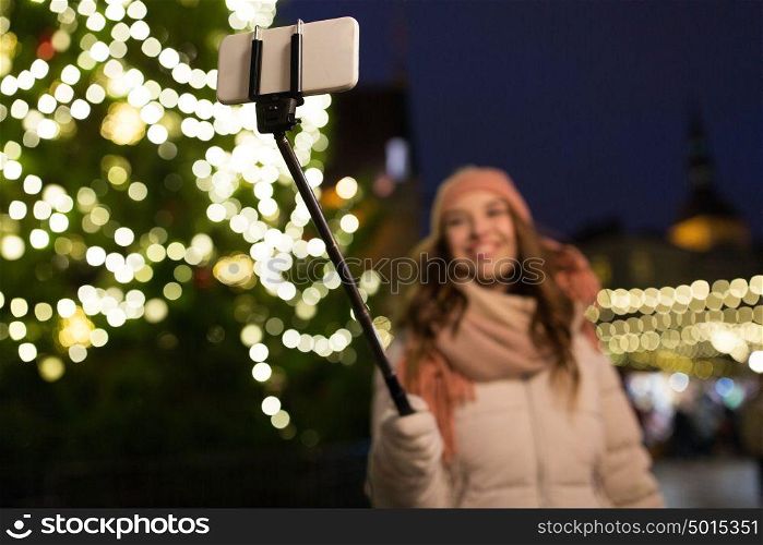 holidays and people concept - beautiful happy young woman taking picture by smartphone on selfie stick at christmas tree and market in winter evening. young woman taking selfie at christmas town