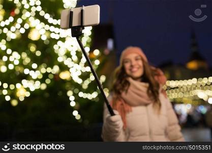 holidays and people concept - beautiful happy young woman taking picture by smartphone on selfie stick at christmas tree and market in winter evening. young woman taking selfie at christmas town
