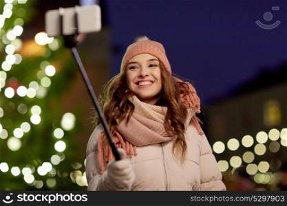 holidays and people concept - beautiful happy young woman taking picture by selfie stick over christmas tree lights in winter evening. young woman taking selfie over christmas tree