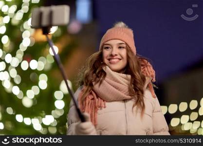 holidays and people concept - beautiful happy young woman taking picture by selfie stick over christmas tree lights in winter evening. young woman taking selfie over christmas tree