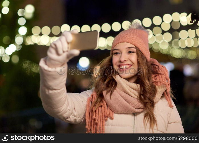 holidays and people concept - beautiful happy young woman taking selfie over christmas lights in winter evening. young woman taking selfie over christmas tree