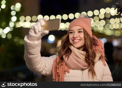 holidays and people concept - beautiful happy young woman taking selfie over christmas lights in winter evening. young woman taking selfie over christmas tree