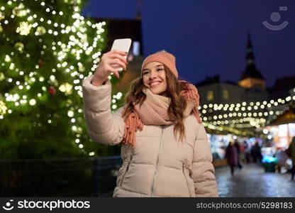 holidays and people concept - beautiful happy young woman taking selfie over christmas tree and market lights in winter evening. young woman taking selfie over christmas tree