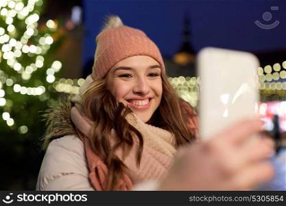 holidays and people concept - beautiful happy young woman taking selfie over christmas tree lights in winter evening. young woman taking selfie over christmas tree